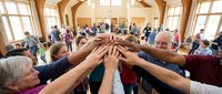 Diverse group of adults joining hands together in a circle during a community gathering in a bright wooden hall, symbolizing unity and teamwork.