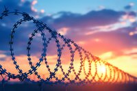 Razor wire silhouette against dramatic sunset sky with orange and blue colors. Security fence detail with barbed spiral creating boundary line at dusk.
