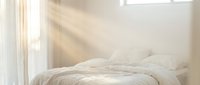 Serene bedroom with unmade bed, soft natural light streaming through window, white linens and curtains creating calm, minimalist aesthetic for relaxation and sleep.