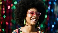 Happy young African American woman with afro hairstyle and retro sunglasses smiling at colorful disco party with bokeh lights background for celebration events.