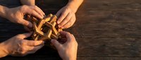 Multiple hands collaborating to assemble a wooden interlocking puzzle on a rustic dark wooden table, symbolizing teamwork and problem-solving.