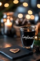 Dark coffee shot glass with white lettering against blurred holiday lights background, pine cones and decorative heart on slate surface creates cozy atmosphere.