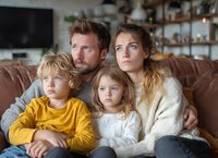 Happy Caucasian family of four sitting together on brown leather sofa in modern living room watching television during cozy evening at home.