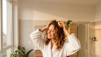 Young woman with curly hair in white bathrobe touching her hair in modern bathroom with natural sunlight streaming through windows for skincare routine.