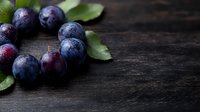 Fresh ripe dark purple plums with water drops and green leaves arranged on black rustic wooden background with copy space for food photography.