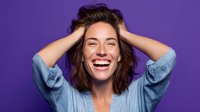 Young Caucasian woman with tousled hair laughing joyfully with hands in hair against purple background, expressing genuine happiness and carefree attitude.