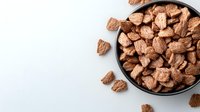 Chocolate cereal flakes in black ceramic bowl on white background, scattered pieces around. Healthy breakfast food concept with copy space.