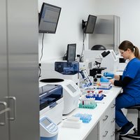 Female scientist in blue lab coat working with microscope and test tubes in modern laboratory conducting medical research and analysis.