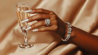 Elegant dark-skinned female hand with long white manicure holding champagne glass, wearing luxury diamond jewelry against silky beige fabric background.
