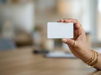 Close-up of hand holding blank white business card indoors with blurred office background. Use for corporate branding, identity design, marketing materials.