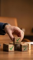 Businessman hand placing wooden blocks with goal checkmarks on desk representing achievement and success planning in warm office environment.