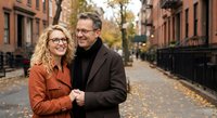 Happy mature Caucasian couple embracing on autumn city street with brownstone buildings. Middle-aged man and woman in coats enjoying romantic walk together.