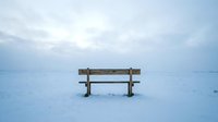 Wooden bench sitting alone on frozen lake covered in snow and ice during winter season with overcast sky.