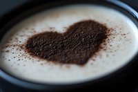 Heart shape made with cocoa powder on top of creamy white coffee drink in dark ceramic cup, romantic coffee art presentation for cafe menu and design.
