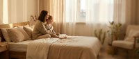 Young Caucasian mother and toddler sit on bed gazing out sunlit window in cozy beige bedroom, warm family moment at home.