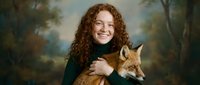 Young woman with curly red hair smiling while holding red fox in autumn forest setting. Wildlife conservation and nature connection concept.