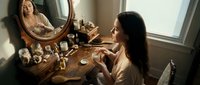 Woman with long dark hair and medium skin tone applying makeup at wooden vanity table with round mirror, cosmetics, and decorative items in bright bedroom setting.