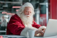Senior man with white beard in red velvet costume working on laptop in modern cafe. Holiday character checking list while enjoying coffee break.