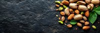 Mixed nuts and pistachios scattered on dark stone background with green leaves and copy space for text, top view panoramic food composition.
