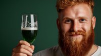 Young caucasian man with red beard and hair holding glass of dark beer with foam against green background, smiling at camera with friendly expression.