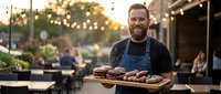 Smiling bearded chef in apron holding wooden tray of grilled meat patties at outdoor restaurant patio with string lights during golden hour sunset.