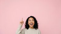 Young woman with dark curly hair and medium skin tone wearing beige sweater, pointing upward with excited expression against pink background for advertising and promotional campaigns.