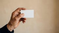 Hand of mature man with medium brown skin tone holding blank white business card against neutral beige background for corporate branding and professional networking purposes.