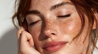 Young woman with freckles and natural makeup enjoying peaceful moment with eyes closed and hand touching face in soft sunlight for skincare and beauty concepts.