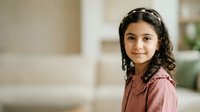 Young girl with long dark hair and South Asian ethnicity wearing a pink button-up shirt, smiling at camera in bright indoor home setting. Portrait for family, lifestyle, and children's content.