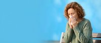 Woman with curly reddish-brown hair, appearing to be in her 40s of Caucasian ethnicity, blowing her nose into a tissue while wearing a sage green sweater against a bright blue background. Suitable for healthcare, wellness, cold and flu, allergy, and medical content.