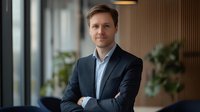Confident male executive in dark suit standing with arms crossed in modern office interior with natural lighting and blurred background for corporate portraits.