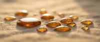 Amber liquid droplets scattered on sandy surface with shallow depth of field, suitable for wellness, skincare, and cosmetic product marketing materials.