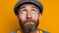 Bearded Caucasian man in knit hat looking up with surprised expression against bright orange background for advertising and marketing campaigns.