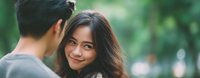 Young Asian woman with long brown hair smiling warmly at Asian man in outdoor park setting with soft natural lighting and green bokeh background.