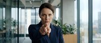 Confident Caucasian businesswoman in navy suit pointing directly at camera with serious expression in modern office environment.