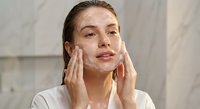 Young Caucasian woman applying white facial cleanser foam to her face with both hands in modern bathroom, skincare routine and beauty treatment concept.