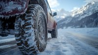 Close-up view of snow-covered off-road tire on winter mountain road with snowy peaks in background, demonstrating extreme weather driving conditions.