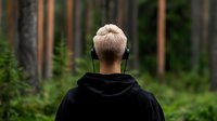 Young person with blonde hair wearing black hoodie and headphones looking at green forest from behind, peaceful solitary moment in nature setting.