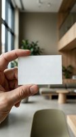 Hand holding blank white business card mockup in modern office interior with natural lighting and wooden furniture for branding presentation.
