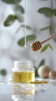Dripping golden honey from wooden dipper into glass jar with blurred green leaves background, natural light setting creates soft reflections on white surface.