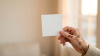 Hand holding blank white card against blurred interior background with natural window light, suitable for business messaging, greeting cards, invitations, and promotional designs.