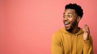 Young African American man wearing mustard yellow hoodie, smiling with excited expression against pink background. Suitable for lifestyle, fashion, and advertising campaigns.