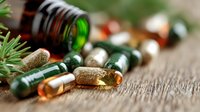 Herbal supplements, vitamins and medicine capsules scattered on wooden surface with green glass bottle and fresh pine sprig, macro view for wellness concept.