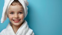 Young girl with bright smile wearing white bathrobe and towel on head against turquoise background, representing spa, wellness and skincare concepts.