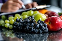 Fresh dark grapes, green grapes and red apple on black reflective surface with water drops. Person writing notes in background, selective focus on fruits.
