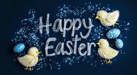 Festive Easter greeting with hand drawn yellow chicks and blue decorated eggs on dark background surrounded by delicate white floral elements and calligraphic text.