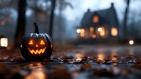 Glowing carved pumpkin on autumn ground with illuminated house in foggy background creates spooky Halloween atmosphere among fallen leaves at dusk.