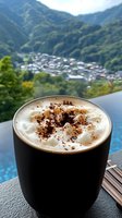 Hot cappuccino with creamy foam and chocolate powder in black matte cup against mountain village backdrop and infinity pool, morning coffee scene.