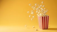 Red and white striped popcorn container with kernels exploding and floating in air against warm yellow background for entertainment and snack concepts.