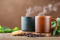 Steaming coffee cups with roasted beans, orange slice and green leaves on wooden surface against brown background, cozy morning atmosphere.